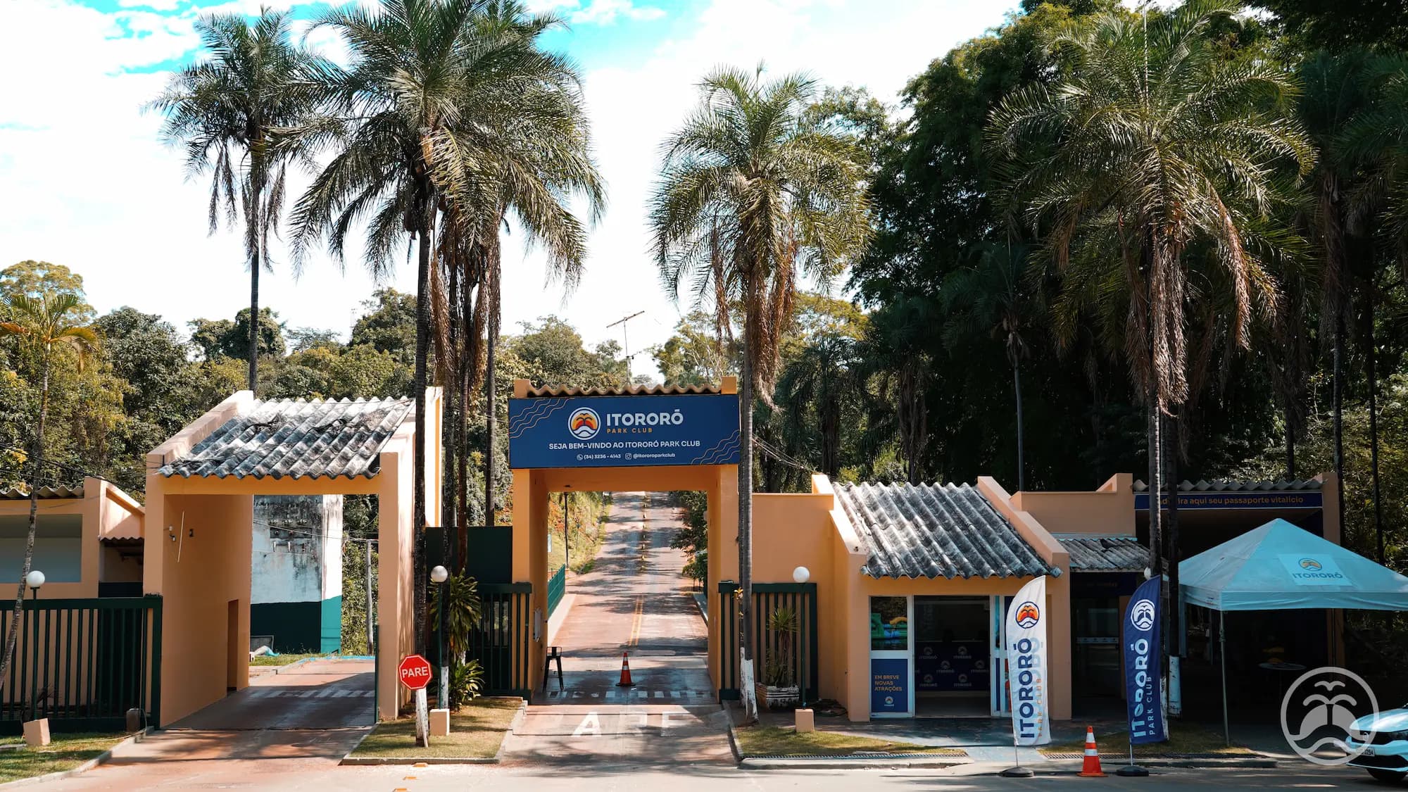 Portaria principal do Itororó Park Club com pista de acesso centralizada e palmeiras ao redor.