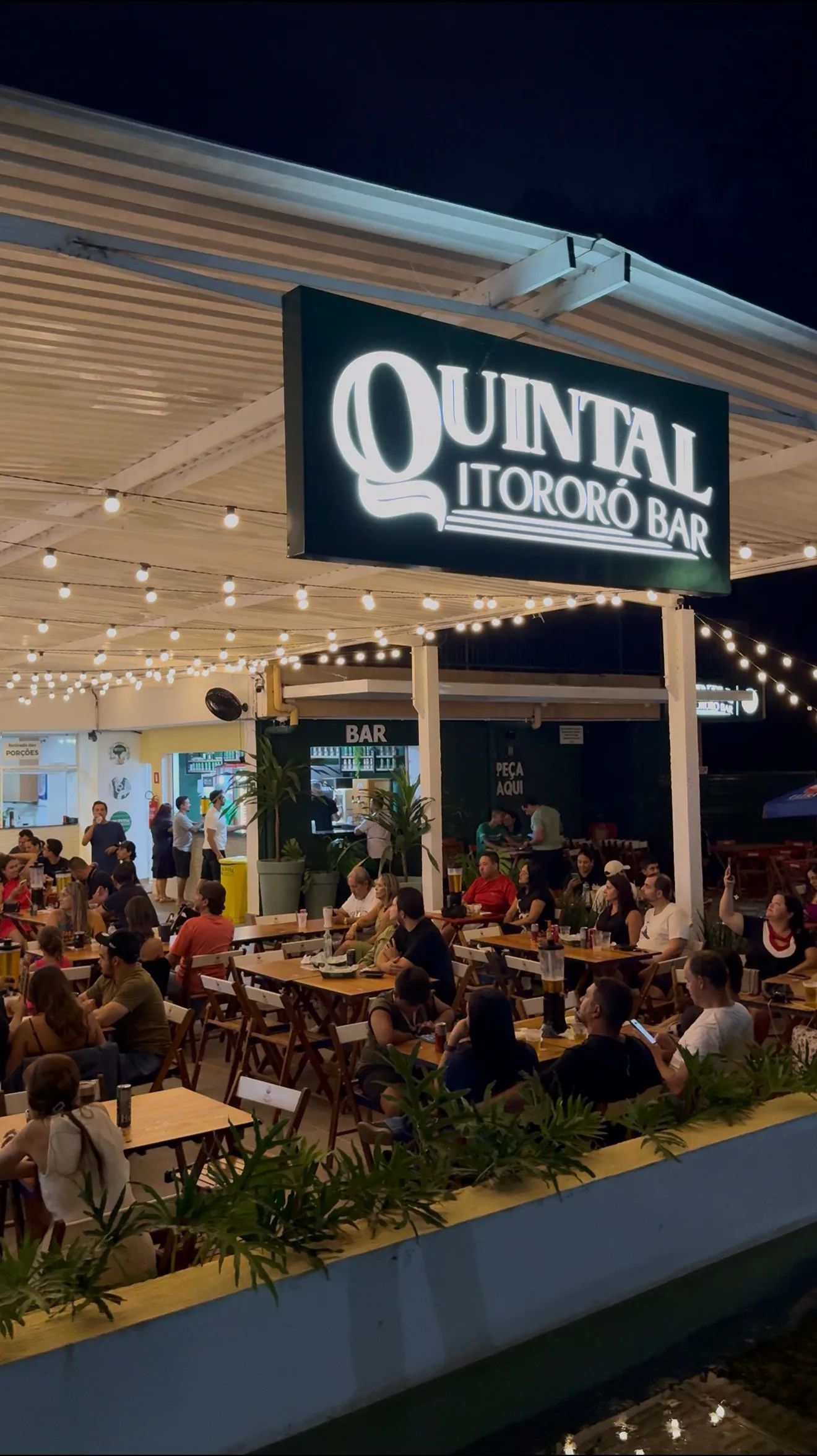Área do Quintal do Itororó Bar com mesas ocupadas e placa iluminada